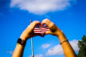 American flag flying in the background with a person making a heart shape with their hands.